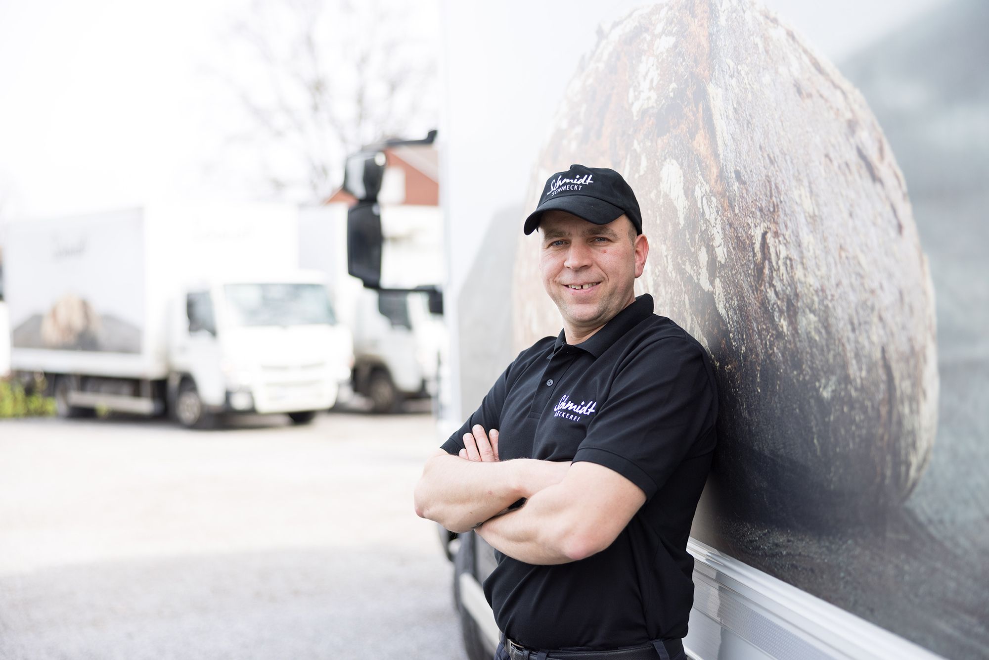 Fahrer der Bäckerei Schmidt steht lächelnd vor einem Lieferfahrzeug mit Brot-Motiv.