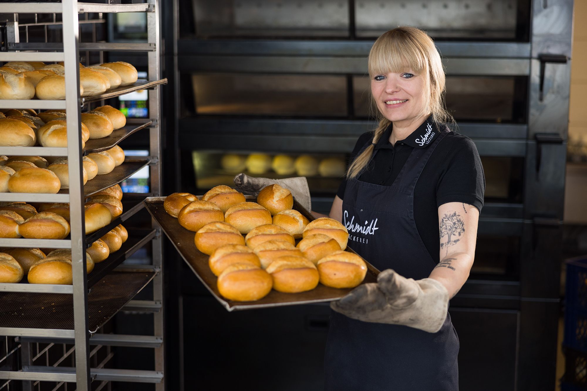 Mitarbeiterin der Bäckerei Schmidt hält ein Blech mit frisch gebackenen Brötchen.