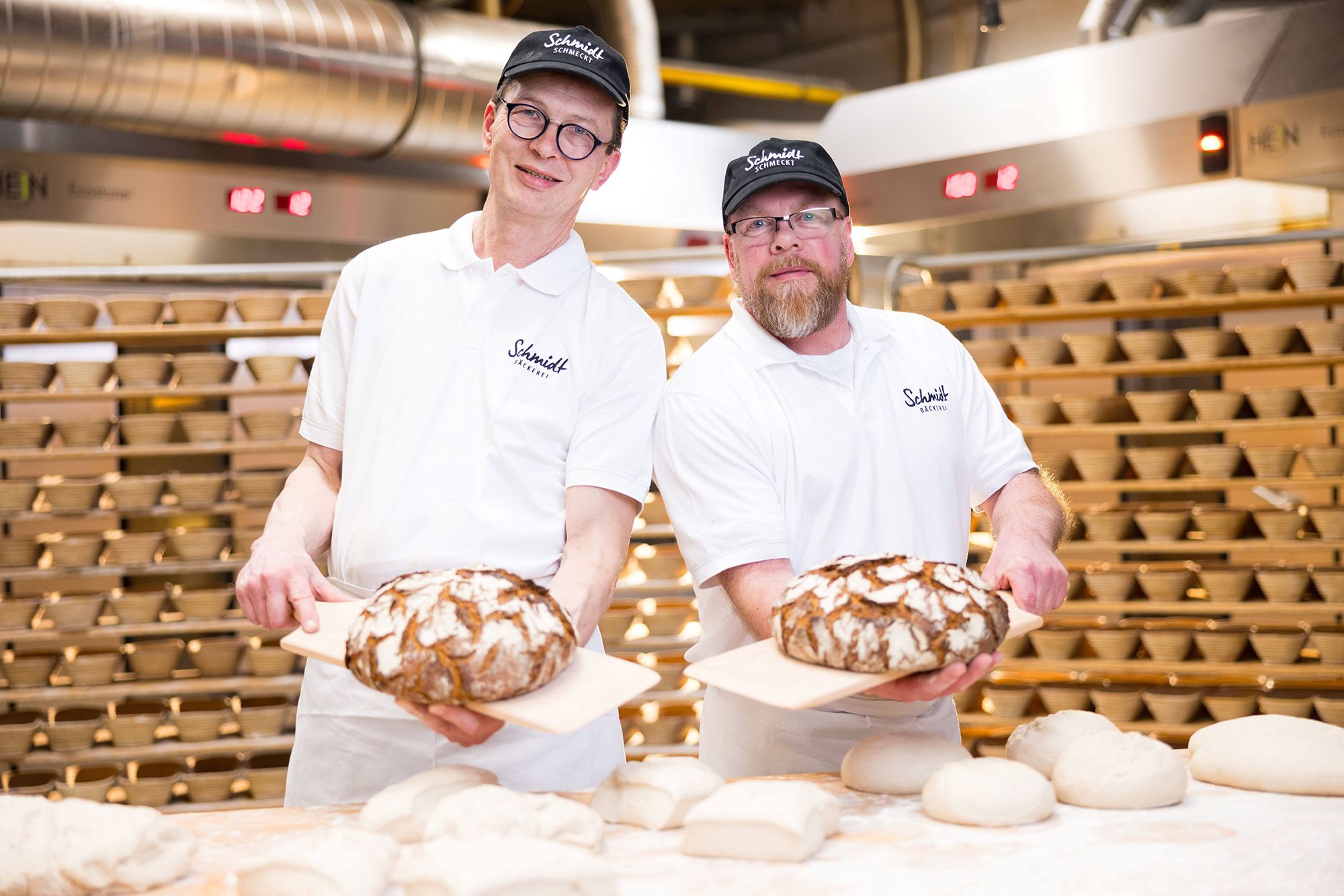 Zwei Bäcker der Bäckerei Schmidt zeigen frisch gebackene Brote in der Backstube.