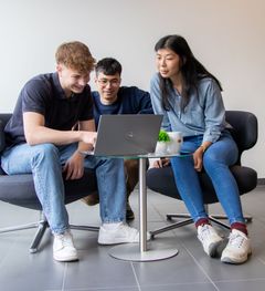 3 junge Kolleg*innen schauen in der Kantine an einem Kaffeetisch gemeinsam in einen Laptop. 