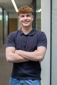Ein junger Mann, er ist Azubi, steht fröhlich lachend im Gang und blickt in die Kamera.
