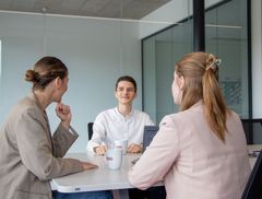 2 junge Frauen sitzen mit dem Rücken zur Kamera und sprechen mit einem jungen Kollegen. Er schaut fröhlich in die Richtung der beiden.