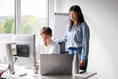 Ein junger Mann sitzt an seinem Arbeitsplatz, er schaut konzentriert in seinen Laptop. Eine junge Kollegin steht dahinter und schau ebenfalls in den Bildschirm.