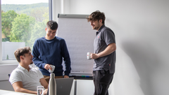 3 junge Männer unterhalten sich an einem Arbeitsplatz. 2 halten Kaffeetassen in der Hand und stehen. der 3. sitzt auf seinem Bürostuhl und hat sich Ihnen zugedreht.
