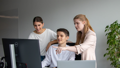 Ein junger Mann sitzt an seinem Arbeitsplatz, 2 Kolleginnen stehen dahinter und zeigen auf seinen Monitor.