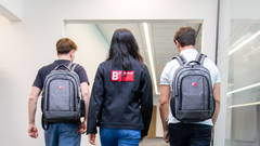 3 junge Mitarbeiter*innen laufen mit dem Rücken zur Kamera durch den Gang. Die Frau in der Mitte trägt  eine Jacke mit Berghof Logo. Die beiden Herren die außen laufen tragen jeweils einen Rucksack