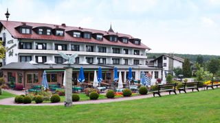 Großes Hotel mit rotem Dach, Sitzbereich mit blauen Sonnenschirmen und Garten davor.