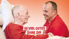 Älterer Mann sitzt im Bett ihm gegenüber sitzt seine Pflegefachkraft. Beide lächeln sich an. So viel Gutes Dank dir.