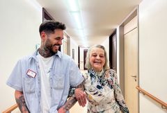 Ältere Dame ist bei der Pflegefachkraft untergehackt. Die Pflegefachkraft lächelt die ältere Dame an. 