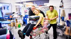 Drei Mitarbeitende des Inklusionsbetrieb. Eine Frau sitzt im Einkaufswagen voller Wäsche und hat Wäsche auf dem Schoß. Zwei Kollegen schieben den Wagen. 