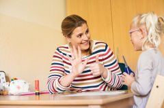 Eine Frau sitzt mit einem kleinen Mädchen am Tisch. Die Frau hält sechs Finger in die Luft und lächelt das Mädchen an. Das Mädchen hält eine Schere in der Hand und lächelt die Frau an. 