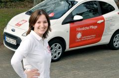 Pflegekraft der Caritas steht lächelnd vor einem Dienstfahrzeug mit roter Beschriftung ‚Häusliche Pflege‘. Sie trägt ein weißes Caritas-Polo und blickt freundlich in die Kamera