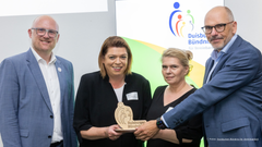Übergabe der Auszeichnung #VerbindlicheVereinbarkeit des Duisburger Bündnisses für Vereinbarkeit. Auf dem Foto sind Anja Rutkat, Sören Link, Michaela Mett und Markus Rüdiger zu sehen. Alle lächeln in die Kamera und stehen vor einer Leinwand. Im HIntergrund ist  eine Präsentationsfolie zu sehen.