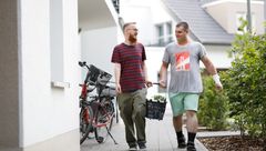 Zwei Männer, lächelnd, tragen gemeinsam einen Einkaufskorb auf einem Gehweg vor Wohnhäusern, mit einem Fahrrad im Vordergrund.