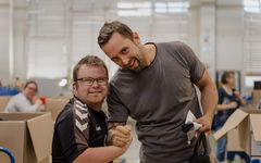 Zwei Männer stehen gemeinsam in der Werkstatt, schauen in die Kamera und halten sich an den Händen fest und lächeln. 