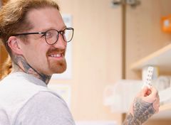 Ein tätowierter, lächelnder Mann mit Brille hält eine Blisterpackung Medikamente vor einem Apothekerschrank.