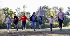 Sieben verschiedene Menschen halten sich an den Händen und laufen gemeinsam die Wiese entlang. Dazu gehören Frauen, Männer und Kinder. Sie alle gehen in die gleiche Richtung.