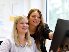 Zwei Frauen schauen auf einen Bildschirm. Die hintere Person zeigt der vorderen etwas auf dem Bildschirm. Beide lächeln.