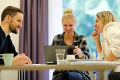 Drei Personen sitzen am Schreibtisch. Die mittlere Person schaut auf ihren Laptop, die anderen beiden schauen sich an. Es ist eine Arbeitssituation, bei der alle drei Personen lächeln.