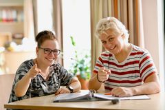 Zwei Frauen sitzen an einem Tisch in einem hellen Raum. Beide lachen herzlich und zeigen mit dem Finger in Richtung Kamera. Vor ihnen liegt ein geöffneter Ordner mit Unterlagen. Die Frau links trägt eine Brille und ein Camouflage-Shirt, die Frau rechts ein gestreiftes Shirt. Die Atmosphäre wirkt freundlich und unterstützend.
