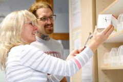 in Mann mit roten Haaren, Bart, Brille und auffälligen Tattoos steht vor einem offenen Regal mit Medikamentenboxen. Er lächelt in die Kamera und hält eine Blisterpackung mit Tabletten in der Hand. Die Szene wirkt freundlich und professionell, vermutlich im Rahmen einer Betreuung oder Pflegeeinrichtung.