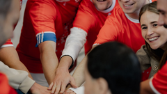 Eine Gruppe von Menschen in roten Trikots mit blauen Akzenten legt ihre Hände übereinander, als Zeichen der Teamarbeit und des Zusammenhalts; eine Frau lächelt im Vordergrund.