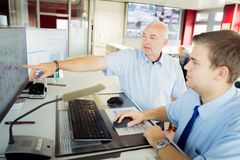Ausbilder erklärt dem Auszubildenden zur Fachkraft im Fahrbetrieb in der Busleitstelle was man alles auf den Monitoren ablesen kann. Im Hintergrund sind zahlreiche Monitore mit den Echtzeitbildern der Kameras von den Busbahnhöfen zu sehen. 