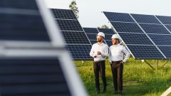 Zwei Männer mit weißen Hemd und weißen Bauhelm schauen in die Ferne; sie stehen zwischen Solarpanels