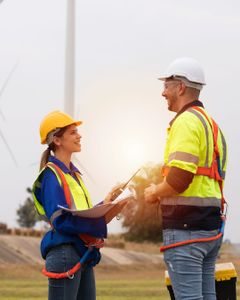 Eine weibliche und eine männliche Person mit Warnwesten und Bauhelm. Im Hintergrund befinden sich Windräder