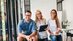 Ein Mann und zwei Frauen, welche in einen Büro sind und freundlich in die Kamera lächeln