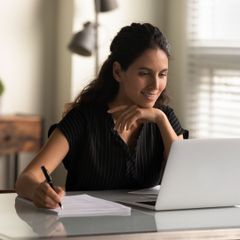 Frau mit Blättern und Stift in der Hand vor einem Laptop
