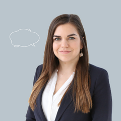 Lächelnde Frau mit langen braunen Haaren, schwarzem Blazer und weißer Bluse vor grauem Hintergrund mit Sprechblasegrauem Hintergrund