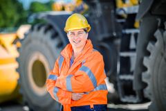 Eine Frau steht mit verschränkten Armen vor einem Radlader. Sie trägt eine orangene Schutzjacke und einen gelben Schutzhelm und lächelt in die Kamera.