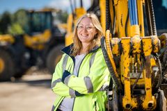 Eine blonde Frau mit Brille steht mit verschränken Armen an einem Baggerarm. Sie trägt eine gelbe Schutzjacke und lächelt in die Kamera.