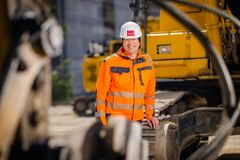 Ein Mann mit einem weißen Schutzhelm und orangener Schutzjacke steht angelehnt an einem Bagger und lächelt in die Kamera.