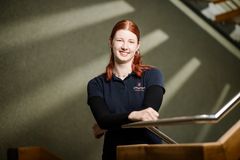 Eine Frau mit roten Haaren, die bei der Stadt Elmshorn arbeitet, lächelt und lehnt an einem Treppengeländer. 