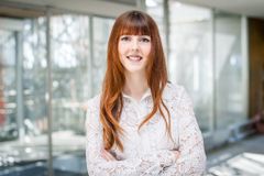 Eine junge Frau  mit roten langen Haaren schaut lächelnd in die Kamera. Sie trägt eine weiße Bluse und im Hintergrund ist eine Fensterfront erkennbar.