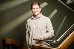 Ein junger blonder Mann mit Oberlippenbart hat einen hellen Pullover an, lächelt in die Kamera und hat eine Hand an einem Treppengeländer.