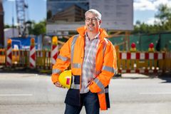 Ein Mann mit Brille trägt eine orangefarbende Schutzjacke. Er trägt unter seinem rechten Arm einen gelben Schutzhelm. Die andere Hand ist in seiner Hosentasche. Im Hintergrund sind Baubarken zu sehen. 