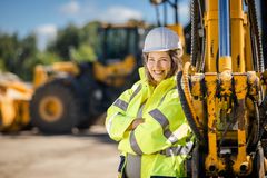 Eine Frau mit gelber Schutzjacke und einem weißen Schutzhelm lehnt an einem gelben Baggerarm und verschränkt die Arme.