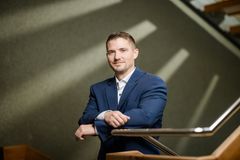 Ein junger Mann mit weißem Hemd im dunkelblauen Anzug lehnt auf einem Treppengeländer und lächelt in die Kamera.