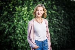 Eine blonde Frau mit halblangen Haaren und der rechen Hand in der Hosentasche  steht vor einer Hecke und lächelt in die Kamera.