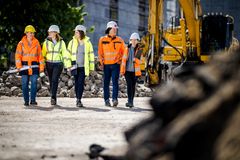 Fünf Personen mit gelber und organgfarbener Schutzkleidung mit Bauhelmen unterhalten sich gehend auf einer Baustelle. Im Hintergrund sieht man graue Wackersteine und einen gelben Bagger.