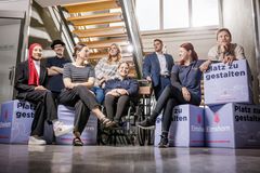 Eine Gruppe von Menschen sitzt im Treppenhaus auf hellgrauen Sitzwürfeln mit dem Schriftzug Platz zu gestalten. Einige schauen lächelnd in die Kamera, andere schauen verträumt in die Ferne.