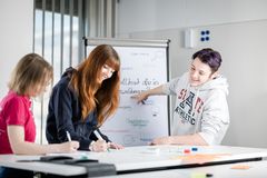 Ein Mann mit liafarbenen Haaren in einem weißen Pullover lehnt am Tisch und zeigt auf ein im Hintergrund sichtbares Flipchart. Zwei junge Frauen schreiben im Vordergrund und lächeln. Die eine Frau hat halblange blonde Haare und ein pinkes Tshirt an, die andere Frau hat lange dunklerote Haare und eine blaue Jacke an.