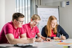 Ein blonder Mann und ein dunkelhaariger Mann in pinken TShirts sitzen mit einer langhaarigen rothaarigen Frau an einem Tisch und schreiben. Im Hintergrund ist ein Flipchart und ein Fenster mit Lamellenvorhängen zu erkennen.