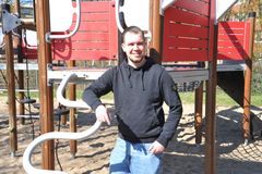 Portrait eines Mannes im blauen Pullover. Er hat kurze blonde Haare und lehnt an einem roten Klettergerüst.