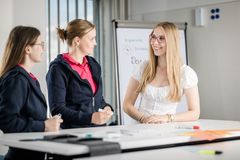 Eine blonde junge Frau mit langen Haaren, Brille und weißer Bluse steht am Tisch und unterhält sich mit zwei zu ihr blickenden  Frauen, die beide ein pinkes Tshirt und eine blaue Jacke anhaben.