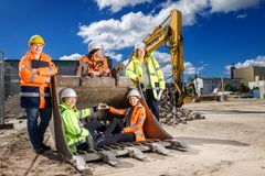 Im Vordergrund einer Baustelle lehnt ein Mann mit Bauhelm sowie zwei Frauen links und rechts mit Bauhelm und Schutzkleidung an einer großen Baggerschaufel. In der Baggerschaufel sitzen zwei Frauen, eine davon mit Brille . Beide haben Schutzjacken an und Bauhelme auf. Die beiden Frauen bilden mit den Händen gemeinsam ein Herz