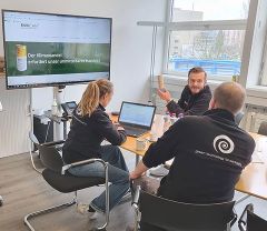 Drei Personen bei einer Besprechung im Büro. Ein großer Monitor zeigt die ENVICAN-Webseite mit der Aufschrift „Der Klimawandel erfordert unser unmittelbares Handeln!